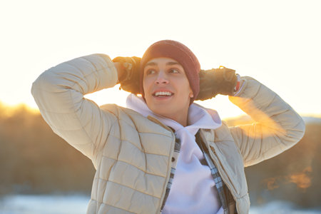 A handsome happy young man in warm clothes stands in a winter park at sunset and admires the beauty...