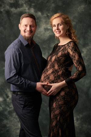A loving husband and a beautiful pregnant wife are expecting a baby. Loving spouses in evening clothes expecting a baby on a grunge studio background.の写真素材