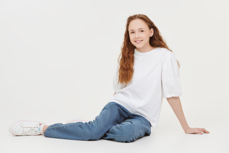 Full-length portrait of a beautiful teenage girl with long wavy hair in a white blouse and blue jeans smiling at the camera. White studio background.の写真素材