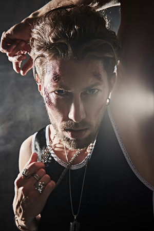 A hardened male bandit with bruises and wounds on his face, who stands in a dark room and looks...