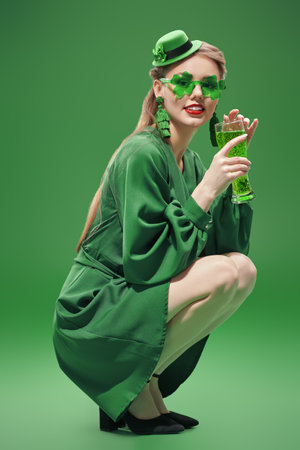 Party for St. Patrick's Day. A cheerful girl, dressed in a green dress, a hat and green clover-shaped glasses, holds a glass of beer. Full-length studio portrait against a green background.の写真素材