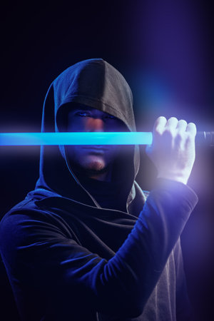 A brave warrior in a black robe with a hood on his head holds a laser sword in front of him on a dark background. Space fantasy. Science fiction novel.の写真素材