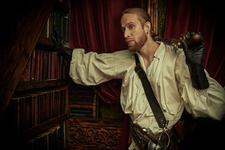 Fantasy. A noble musketeer with a rapier stands in the library. Late Middle Ages, 16th-17th century. The hero of a historical novel.の写真素材
