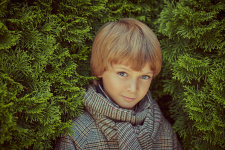 Portrait of a little boy in a checkered coat and scarf standing among green bushes. Children's fashion.の写真素材