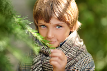 A dreamy little boy in warm checkered clothes stands among green bushes and looks at the falling snowflakes. Winter day with snowfall. Kid's fashion and emotions.の写真素材