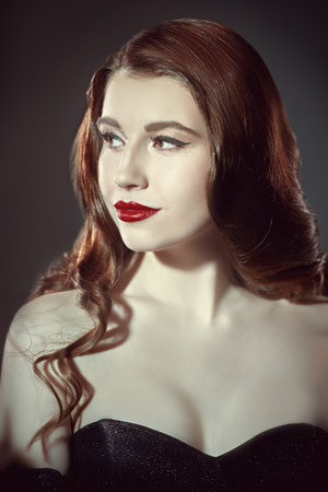 Evening hair and makeup. Portrait of a beautiful girl with long wavy hair and bright evening make-up with red lips, posing in an evening black dress with a neckline. Studio shot.の写真素材