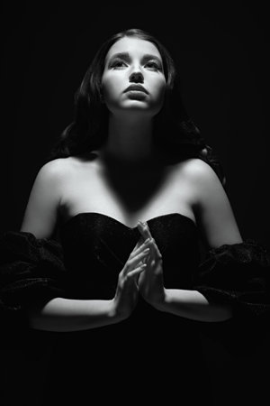 Black and white portrait of a beautiful lady with black wavy hair and elegant makeup with arrows standing in a black low-cut dress in a beam of light on a black background. Retro Hollywood style.の写真素材