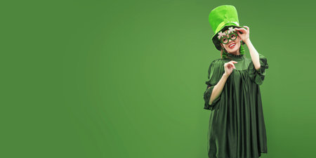 St.Patrick 's Day. Cheerful attractive girl dressed in a green dress, leprechaun hat and green clover-shaped glasses is smiling. Studio portrait on a green background with copy space.の写真素材