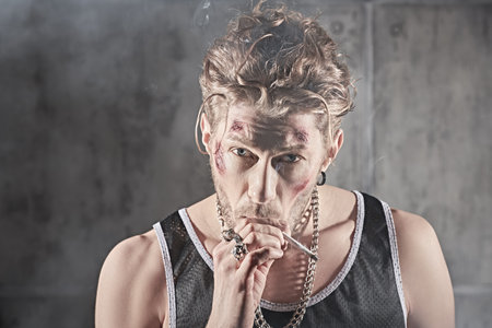 A shot of a hardened male bandit with bruises and wounds on his face, who stands in a dark room with concrete walls and smokes a cigarette. Bad guy concept. Action movie hero.の写真素材