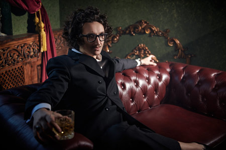 An imposing man with a stylish curly hairstyle in a classic suit and glasses drinks whiskey while...