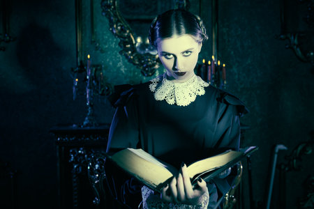 The Addams Family novel. A serious brunette girl in a neat black dress with a white collar stands in the night in a gloomy gothic castle and reading an ancient book. Mystical novel, gothic.の写真素材