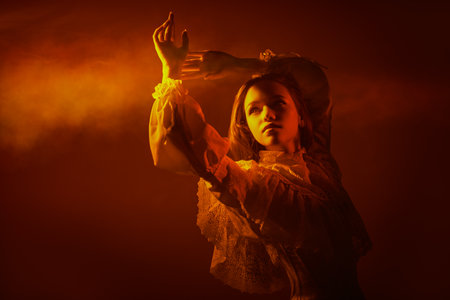 Portrait of a beautiful romantic girl in a long white lace dress, who looks at a beam of light, shrouded in haze. Art portrait. Studio shot on a dark red background. Copy space.の写真素材