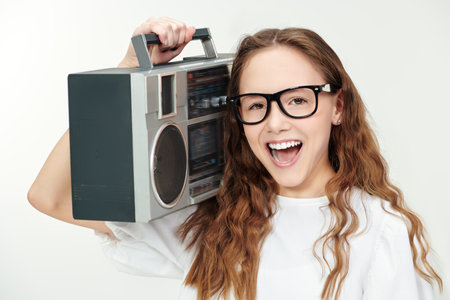 Shot of a beautiful happy teenage girl with long wavy hair in a white blouse, glasses and holding a stereo recorder. White studio background. Music.の写真素材