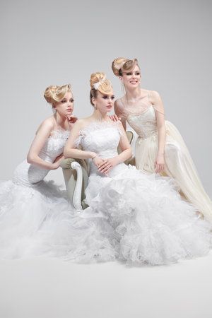 Three elegant blonde brides in puffy white dresses pose in the studio on a grey background. Extravagant hair and makeup. Wedding fashion.の写真素材