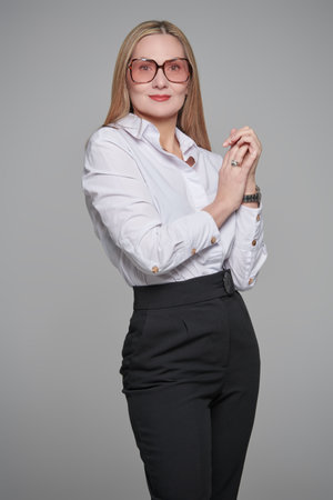 Business fashion. Portrait of a happy beautiful middle aged businesswoman in a classic formal outfit and spectacles looking at the camera and smiling. Glasses style. Grey studio background.の写真素材
