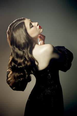 Retro Hollywood style. Elegant evening makeup and hairstyle. Portrait of a charming young woman in a luxury black dress on a studio background.の写真素材