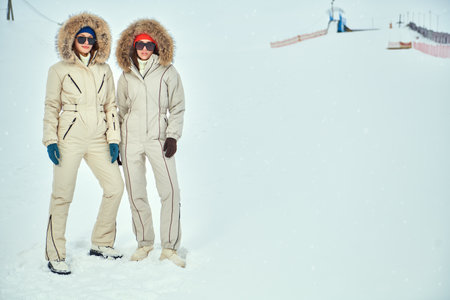 Alpine skiing, active winter recreation. Winter fashion. Full-length portrait of two fashion model girls posing in white downy overalls against the backdrop of a snowy landscape. Copy space.の写真素材