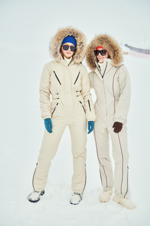 Alpine skiing, active winter recreation. Full-length portrait of two fashion model girls posing in white downy overalls against the backdrop of a snowy landscape. Winter fashion.の写真素材