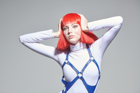 Stylish cyberpunk girl with bright red makeup and red hair posing in white overalls against a grey studio background. Virtual games. Makeup and hairstyle.の写真素材