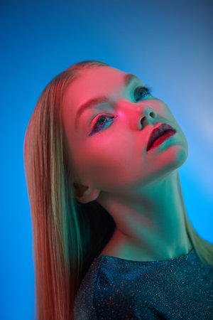Fashion and style with glitter. Stunning blonde girl with shiny glitter makeup and a sparkling evening dress poses on a blue background in mixed colored light. Hairstyle.の写真素材