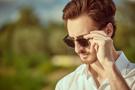 A well-groomed handsome confident man in elegant black sunglasses and a white shirt stands outdoors on a sunny day. Male beauty and fashion. Summer vacation. Copy space.の写真素材
