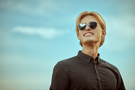 Eyewear fashion. An attractive male model with blond hair posing in black shirt and stylish sunglasses against the sky background. Copy space.の写真素材