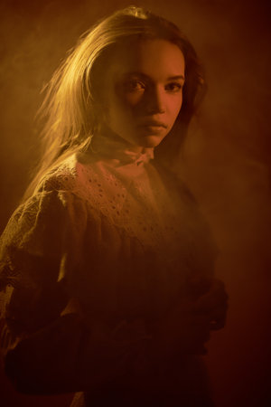 Portrait of a beautiful romantic girl wearing old-fashioned white lace dress, standing alone in a room  shrouded in haze. Art portrait in vintage style.の写真素材