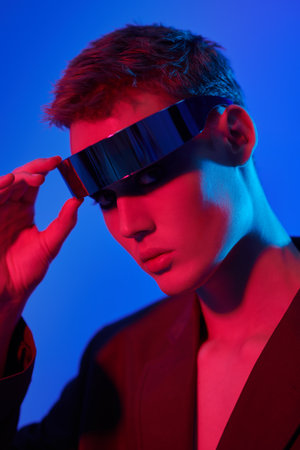 Fashion concept. Portrait of a stylish young man posing in mirrored futuristic glasses in mixed neon blue and crimson light. Blue studio background. Glasses style.の写真素材