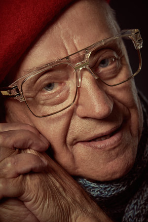 Portrait of an intelligent elderly man in a beret, scarf and glasses standing with folded hands next to his face and looking with a calm smile into the camera. Dark background. Old age concept.の写真素材