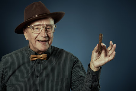 An old gentleman smokes a cigar with pleasure and smiles. Detective story character. Studio portrait on a dark blue background.の写真素材