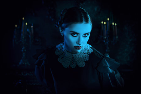 Gothic black comedy. A serious brunette girl in a neat black dress with a white collar stands in the night in a gloomy gothic castle and looks unfriendly to the camera. The Addams Family.の写真素材