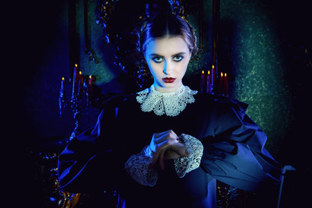 An unfriendly brunette girl in a neat black dress with a white collar stands in the night in a gloomy gothic castle and looks to the camera. The Addams Family. Gothic black comedy.の写真素材