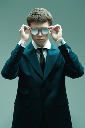 Glasses style. Handsome well-groomed young man posing in a classic white shirt, elegant suit and stylish round sunglasses on a grey studio background. Men's fashion, classic style and beauty.の写真素材