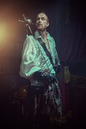 Portrait of a handsome brave musketeer in a white shirt standing in a dark vintage room and holding a rapier in his hands. 16-17 centuries. Historical novel. The era of romanticism.の写真素材