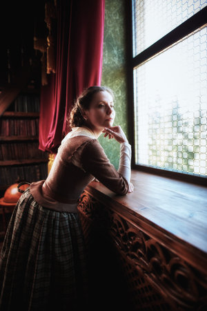A beautiful aristocratic girl in an 18th century Scottish dress looks thoughtfully out the window in...