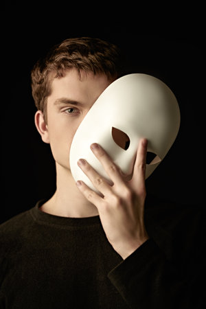 An emotionless young man in a black pullover holds a white mask in his hand, covering part of his face with it. Black studio background. Roles of people. Psychology, inner world.の写真素材