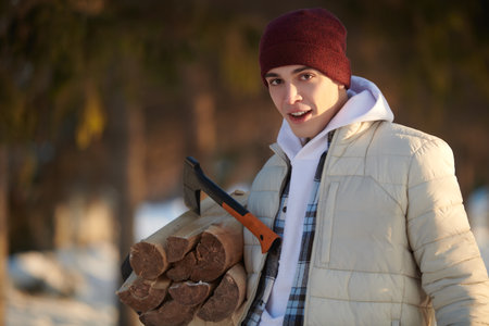 Active recreation in winter, travel and tourism. A young man in warm winter clothes holds an armful of firewood, standing in a winter snowy forest.の写真素材