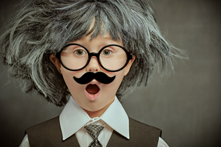 Funny cute kid in glasses, with a mustache and in a wig, makes a surprised face. Against the background of a black chalkboard. Little Scientists. Education and early development.の写真素材