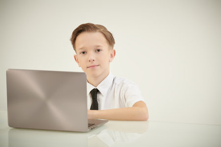 Modern education of children. A smart boy schoolboy in a white shirt and a tie sits at a laptop and smiles. White background with copy space.の写真素材