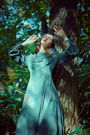 The heroine of a fairy tale. A beautiful brunette girl in a long dress stands by a tree in a magical forest in warm rays of the sun. World of fantasy.の写真素材