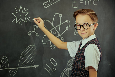 Portrait of a smart boy in a school uniform and glasses looking at the camera seriously and smiling. The background with a black chalkboard with scientific drawings. Education. Young scientists.の写真素材