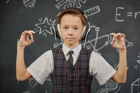 Portrait of a smart boy in a school uniform and headphones looking at the camera calmly. The background with a black chalkboard with scientific drawings. Education. Young scientists.の写真素材