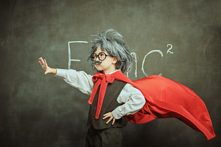 Education and early development. Little Scientists. Cute little kid in glasses, with a mustache and in a wig, like little Einstein makes a pose of superhero. Background of a black chalkboard with copy space.の写真素材
