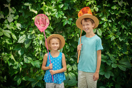 Two happy children stand among green foliage with butterfly nets. Joyful summertime. Happy summer holidays, children's games. Active kids.の写真素材
