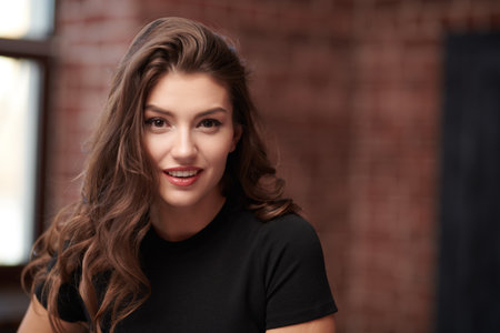 Attractive confident young woman with beautiful long curly hair posing in a loft room in a black top. Beauty and fashion. Confident lifestyle.の写真素材