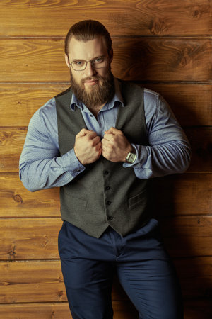 Handsome well-groomed male gentleman in a classic respectable suit with a vest and shirt on a wooden background. Men's hairstyles and beard. Male beauty and fashion. Successful businessman.の写真素材