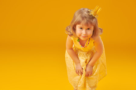 Cute little girl in a princess dress and a crown. Pretty child preparing for a costume party or birthday. Kid's fashion. Portrait on a studio yellow background with space for text.の写真素材