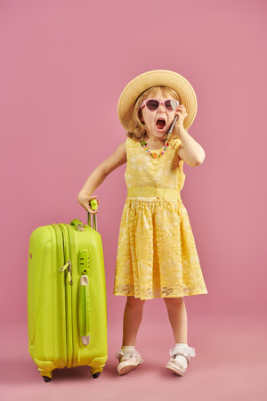 A funny little girl in a fashionable yellow dress and hat stands next to her suitcase and screams loudly into the phone. Summer vacation. Tourism. Studio portrait on a pink background. Place for text.の写真素材