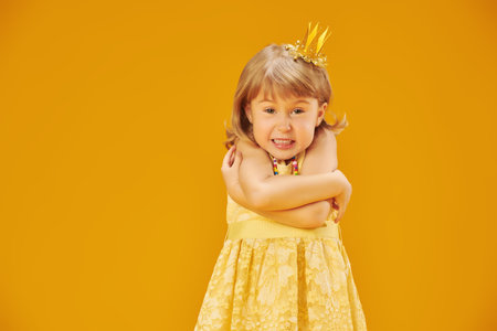 A little capricious girl princess in a beautiful yellow dress and a golden crown is very angry. Children's emotions. Studio yellow background with space for text.の写真素材