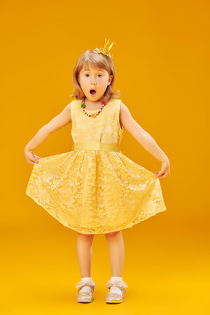 A beautiful cute little princess in a festive yellow dress and a golden crown is very surprised. Full length portrait on a yellow studio background. Fashion for children.の写真素材
